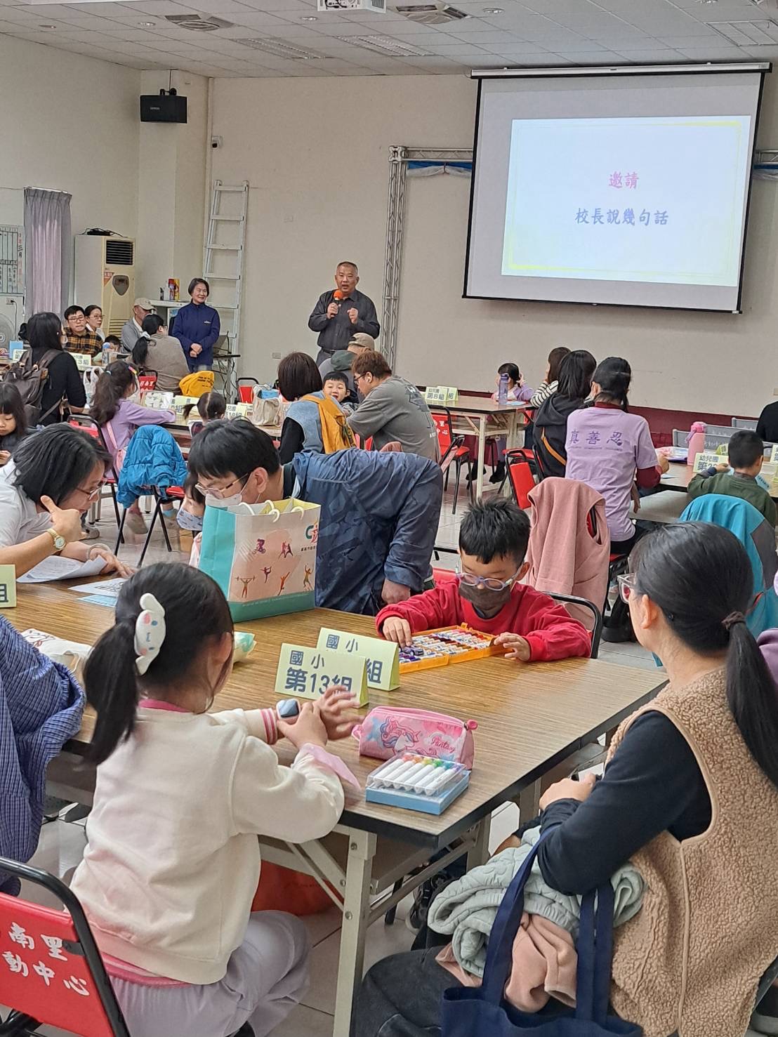 社區著色畫比賽現場 1 — 親子於各組桌位專注著色，校長受邀上台致詞